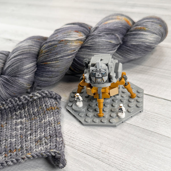 knit swatch and a skein of hand dyed wool yarn for knitting and crochet in shades of gray and gold speckles lunar lander space color theme with lego lunar lander saturn 5 